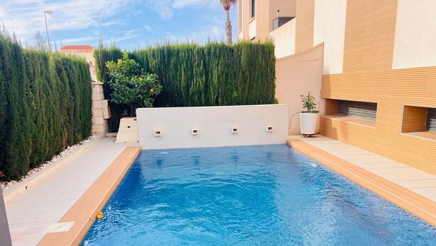 A rectangular swimming pool with a tiled deck, surrounded by a low hedge and a building with a beige facade.