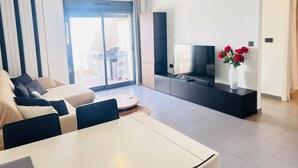 A modern living room with a sofa, a dining table, a TV on a black stand, and a vase with red flowers.