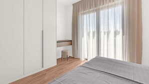 Ein Schlafzimmer mit Bett, Schrank, einem kleinen Tisch und einem Fenster mit durchsichtigen Vorhängen.