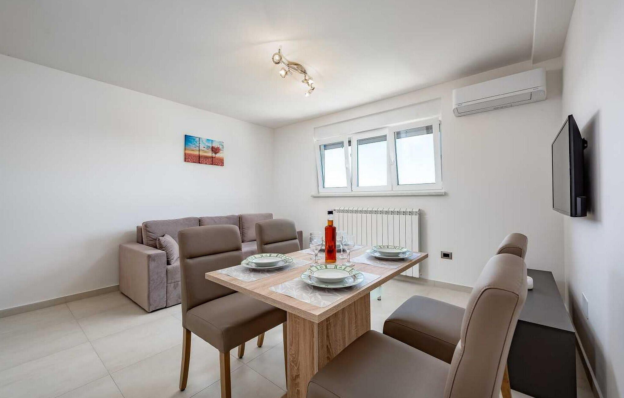 A modern living room with a wooden dining table, beige sofa, and a flat-screen TV mounted on the wall.