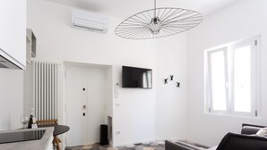 Una sala de estar moderna y bien iluminada, con un sofá, una mesa de comedor con sillas, un televisor montado en la pared y una lámpara de techo original.