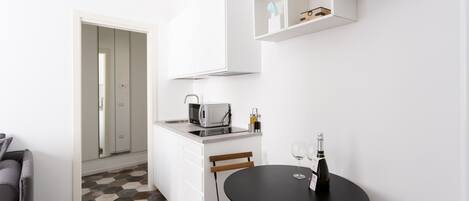 A compact kitchen with a microwave, sink, and a small dining table with two chairs. There is a potted plant on a shelf above the counter.