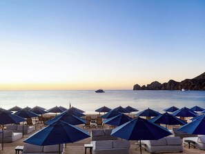 Un hotel frente a la playa con filas de tumbonas y sombrillas, un barco en el agua y acantilados rocosos al fondo.