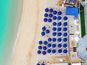 Vista aérea de um hotel à beira-mar com fileiras de guarda-sóis azuis e cadeiras de praia brancas.