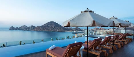 Zona junto a la piscina con sillones reclinables y sombrillas, con vista a un cuerpo de agua y un horizonte urbano distante.