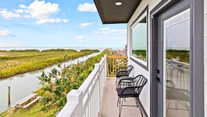 A balcony with a view of a river and a bridge, featuring a wooden picnic table and black chairs. - Experience coastal luxury at Sunny Isle Sanctuary. This chic 2BR/2BA features a designer kitchen, private bay-view balcony, and modern finishes. Reliable service meets the comfort of home. Perfect for your high-end SPI getaway. Book your stay now! (South Padre Island)
