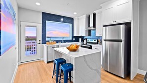 A modern kitchen with a large island, stainless steel appliances, and a view of the sunset through a window. - Experience coastal luxury at Sunny Isle Sanctuary. This chic 2BR/2BA features a designer kitchen, private bay-view balcony, and modern finishes. Reliable service meets the comfort of home. Perfect for your high-end SPI getaway. Book your stay now! (South Padre Island)