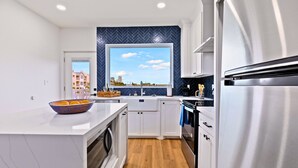 A modern kitchen with white cabinets, a stainless steel refrigerator, and a large window overlooking a cityscape. - Experience coastal luxury at Sunny Isle Sanctuary. This chic 2BR/2BA features a designer kitchen, private bay-view balcony, and modern finishes. Reliable service meets the comfort of home. Perfect for your high-end SPI getaway. Book your stay now! (South Padre Island)