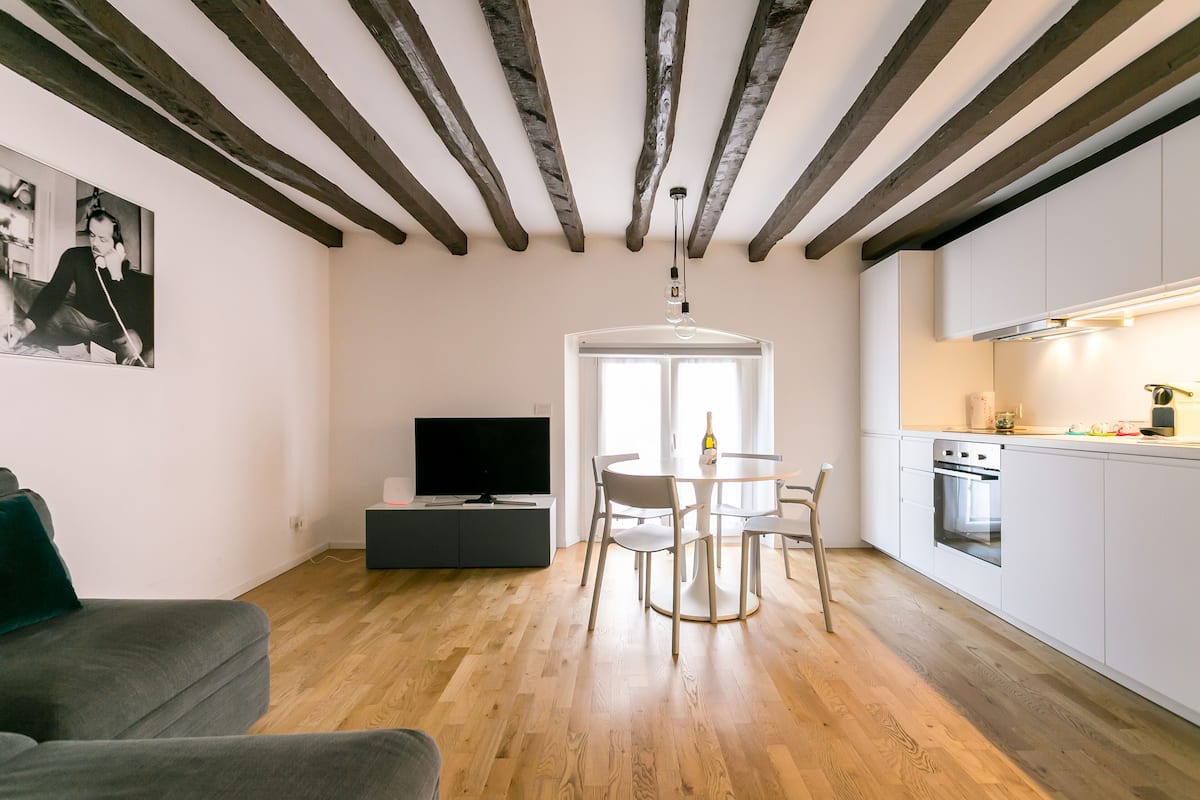 A modern living room with a wooden floor, a grey sofa, a dining area with a table and chairs, a kitchen with white cabinets and a built-in oven, and a ceiling with exposed beams.