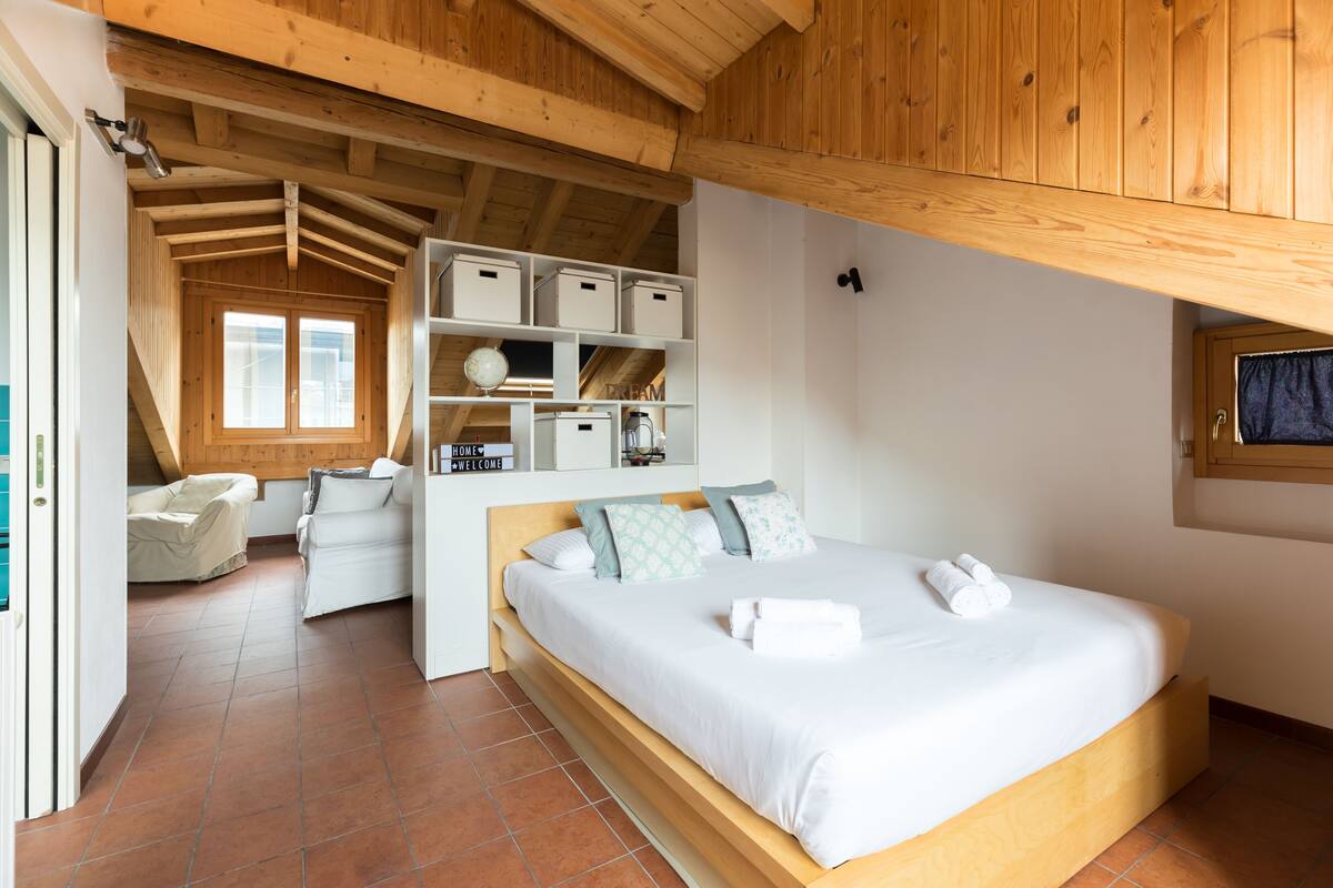 A bedroom with a wooden ceiling, a bed with white bedding, a small seating area with a chair and sofa, and a window with a view.