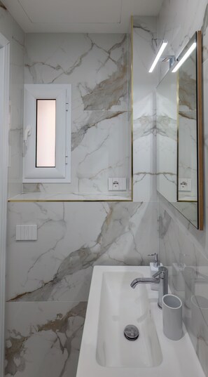 A bathroom with a marble wall, a rectangular mirror, a sink with a modern faucet, and a light fixture above the mirror. - Garibaldi 125 (Naples)