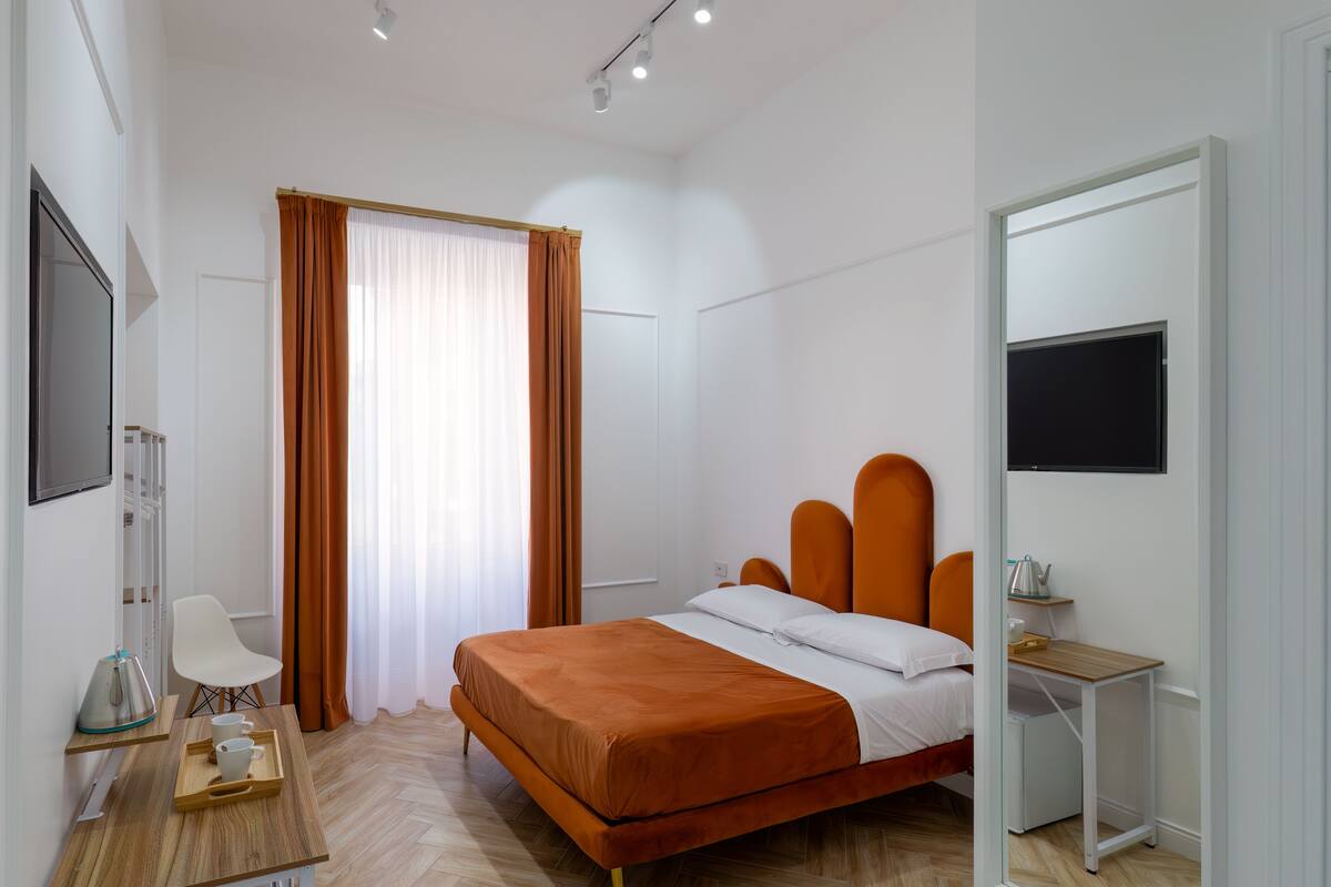 A modern bedroom with a large bed, a desk with a tray of cups, a chair, and a small table with a kettle.