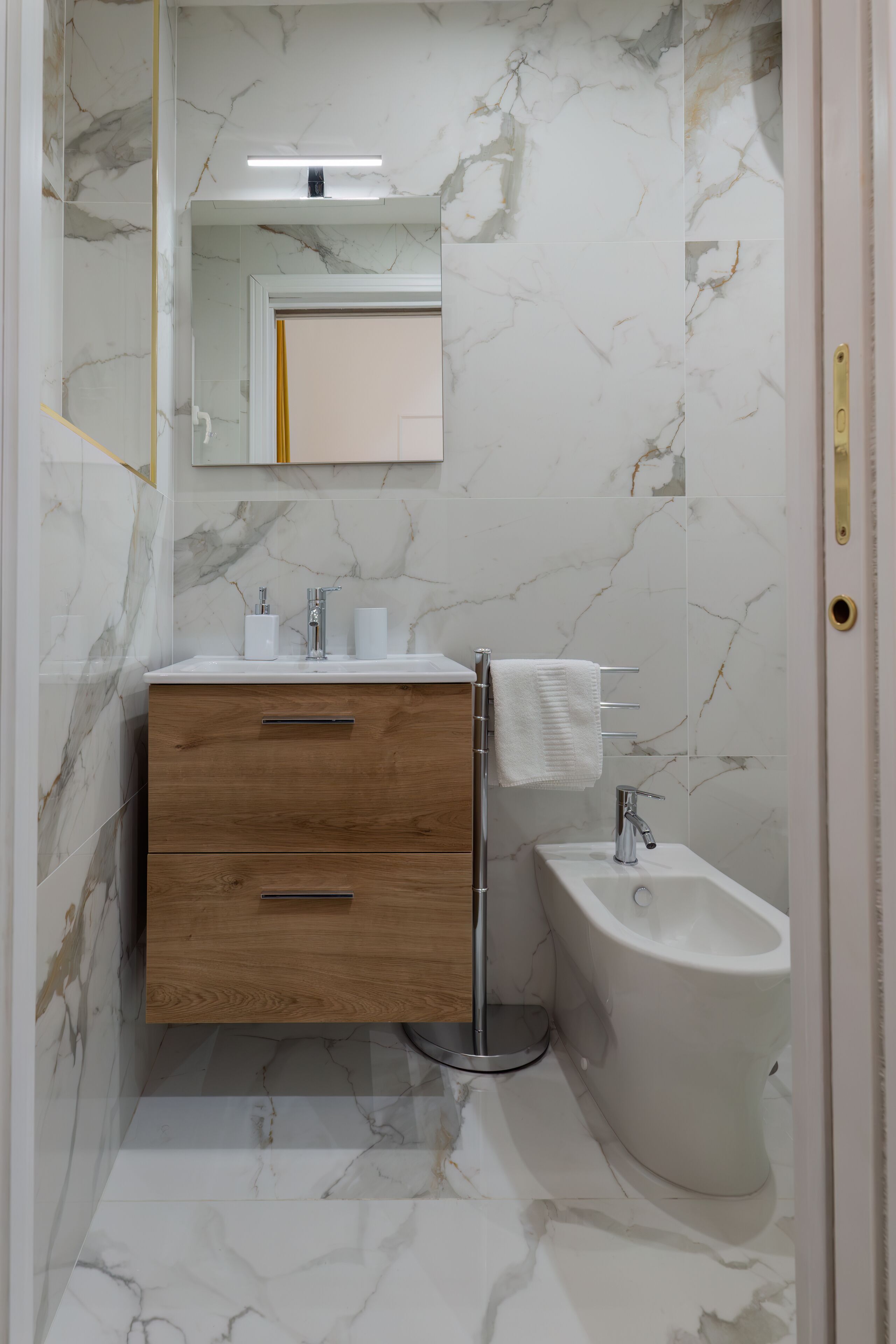Un baño con pared de mármol, tocador de madera, lavamanos, bidé y espejo.