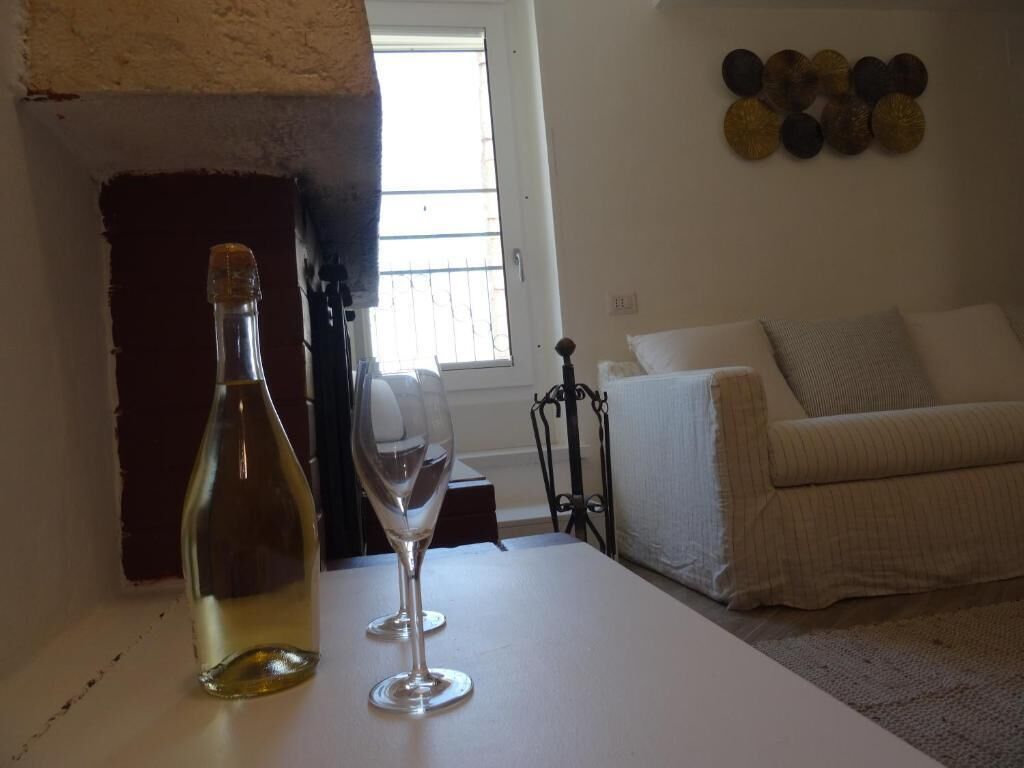 A room with a white table, a bottle, and a wine glass. There is a window with blinds, a white couch, and a wall with decorative plates.