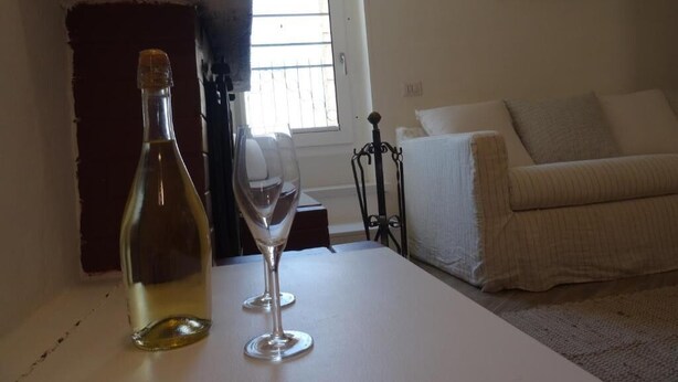 A room with a white table, a bottle, and a wine glass. There is a window with blinds, a white couch, and a wall with decorative plates. - LA DIMORA DELLE 3 ZIE (Matera)