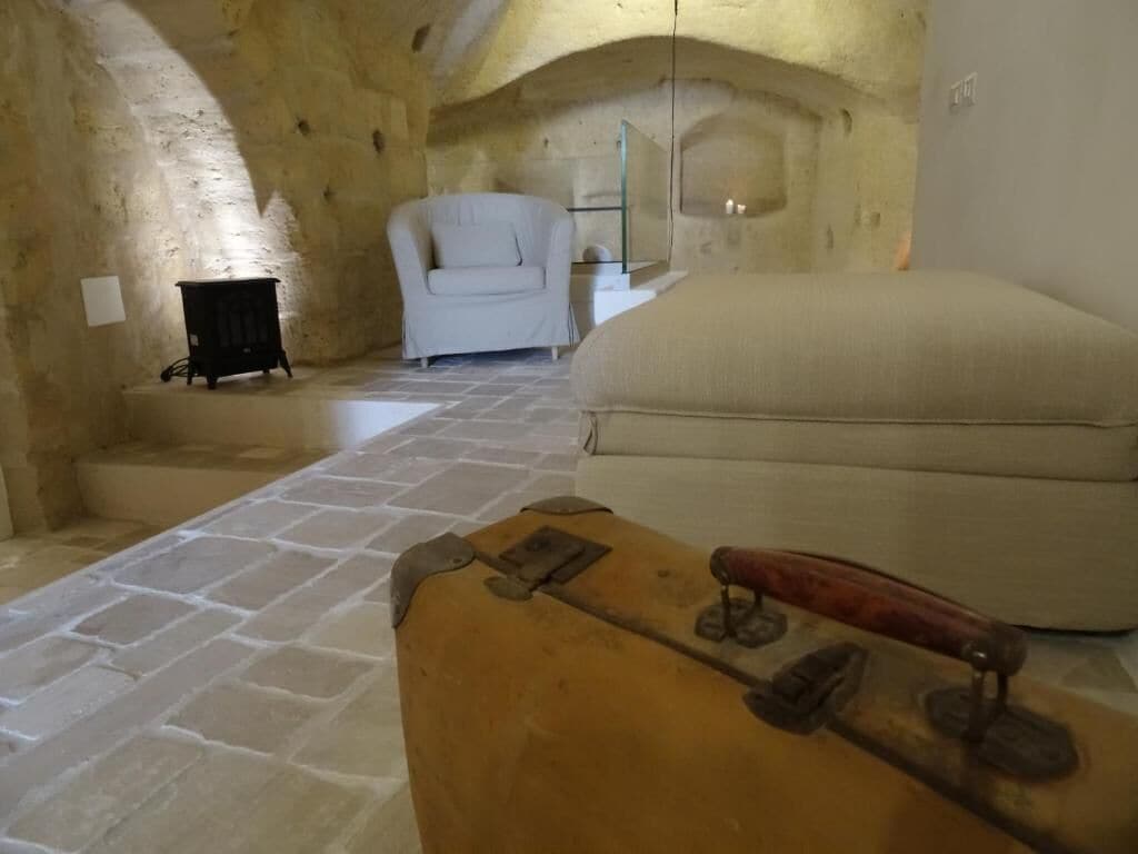 A stone-walled room with a large, round, beige sofa and a white armchair. A wooden chest with metal fittings is in the foreground.