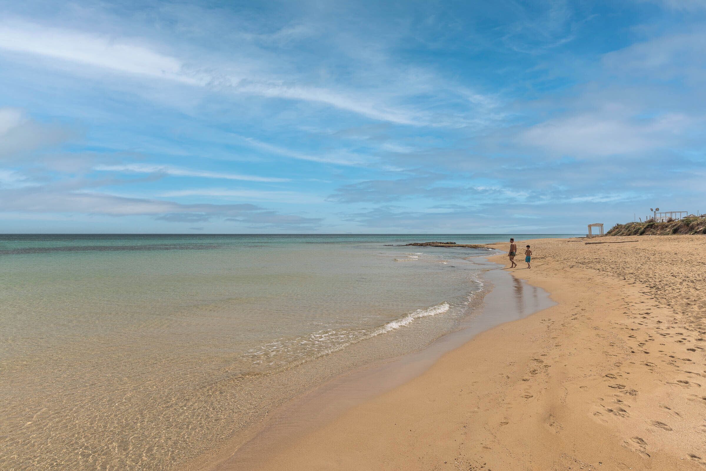 Une plage à l’eau claire, avec un rivage sablonneux, où quelques personnes se promènent.