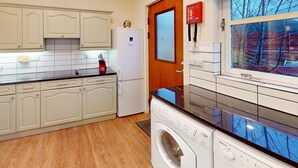 A compact kitchen with white cabinets, a built-in washing machine, and a window with a view of trees. - Pine Marten Cottage Aviemore  | Cosy Holiday Cottage in Silverglades (Aviemore)