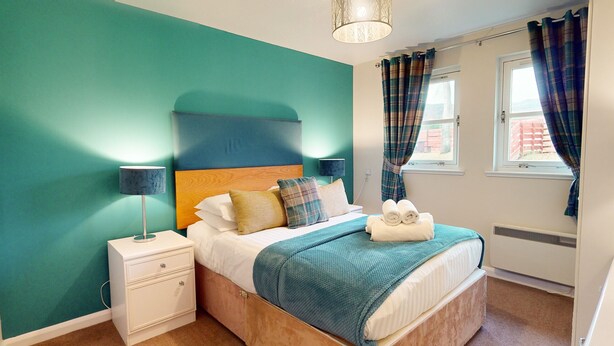 A bedroom with teal walls, a wooden headboard, a bed with white bedding, a bedside table with a lamp, and plaid curtains. - Pine Marten Cottage Aviemore  | Cosy Holiday Cottage in Silverglades (Aviemore)