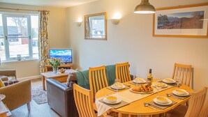 A dining area with a round table set for four, surrounded by wooden chairs. There is a television on a stand to the left, and a window with curtains to the left of the table. - Pine Marten Cottage Aviemore  | Cosy Holiday Cottage in Silverglades (Aviemore)