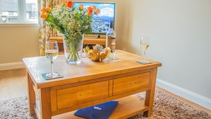 A wooden coffee table with a vase of flowers, two glasses of wine, and a bowl of oranges. A television is in the background. - Pine Marten Cottage Aviemore  | Cosy Holiday Cottage in Silverglades (Aviemore)