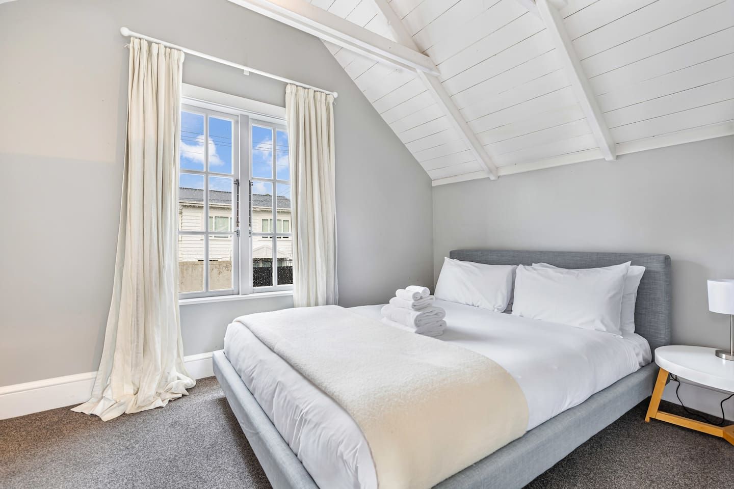 Ein Schlafzimmer mit einem Bett, einem Nachttisch, einem Fenster mit Vorhängen und einer geneigten Decke.