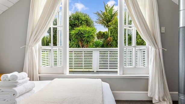 Un dormitorio con un ventanal grande, cortinas blancas, una cama con ropa de cama blanca y vistas a la vegetación exterior.