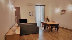 A modern living room with a sofa, a TV, a dining table with chairs, and a decorative wall piece. - Athena Apartment Ortigia Holidays (Siracusa)