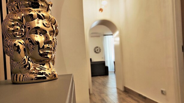 A hallway with a golden sculpture, wooden flooring, and a clock on the wall. - Athena Apartment Ortigia Holidays (Siracusa)