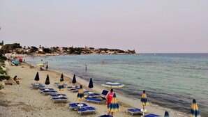A beach with rows of sun loungers and umbrellas, a calm sea, and a distant shoreline with buildings. - Perla del Golfo Ortigia Holidays (Fontane Bianche)