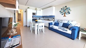 A modern living room with a blue sofa, white dining table, and a kitchen in the background. - Perla del Golfo Ortigia Holidays (Fontane Bianche)
