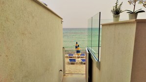 A view from a balcony to a beach with a sandy area, a blue and yellow umbrella, and a potted plant. - Perla del Golfo Ortigia Holidays (Fontane Bianche)