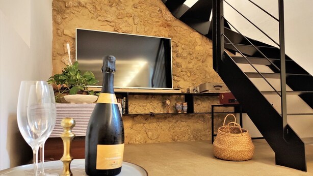 Un salon moderne avec un mur en pierre, un téléviseur à écran plat fixé au-dessus d’une cheminée, une bouteille de champagne, un verre et un sac en fibres naturelles posés sur une table ronde.