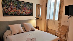 Un lit bien fait, avec deux oreillers et des serviettes enroulées, un tableau au-dessus du lit, une chaise et un téléviseur fixé au mur.