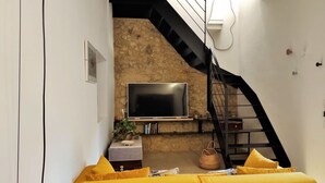 Un salon avec un canapé jaune, un téléviseur à écran plat fixé à un mur en pierre et un escalier noir.