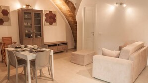 A dining area with a white table and chairs, a cabinet with glass doors, and a sofa in a room with a sloped ceiling and a door. - Domus 1333 Ortigia Holidays (Siracusa)