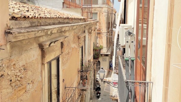 A narrow urban street with buildings on both sides, featuring balconies and a narrow alleyway.