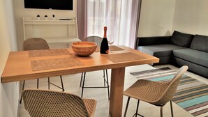 A modern living room with a wooden dining table, wicker chairs, a flat-screen TV mounted on the wall, and a sofa.