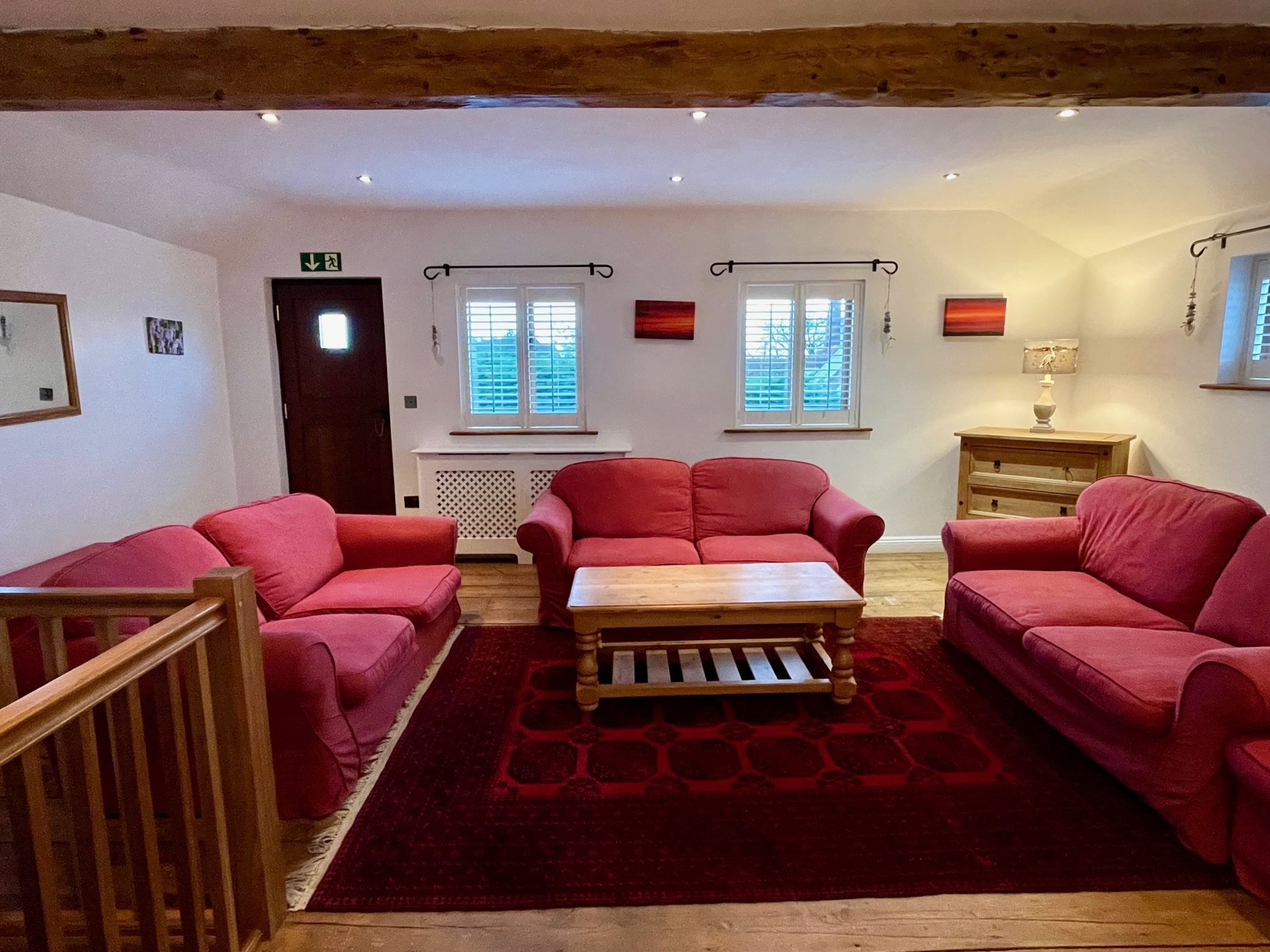 A cozy living room with red sofas, a wooden coffee table, and a red rug. There are two windows with blinds, a door, and a small table with a lamp.