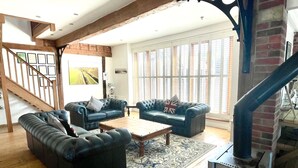 A spacious living room with a wooden staircase, a fireplace, and large windows with shutters. - The Cart Lodge at Lee Wick Farm Cottages (Clacton on Sea)