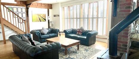 A spacious living room with a wooden staircase, a fireplace, and large windows with shutters.