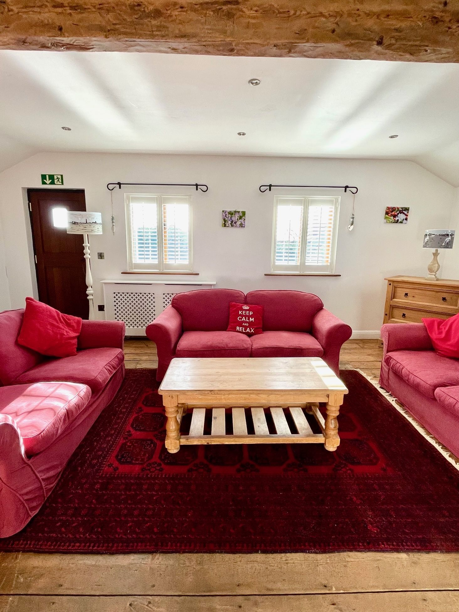 Une pièce chaleureuse avec un canapé rouge, une table basse en bois et un tapis rouge. On y trouve deux fenêtres équipées de stores, un radiateur et un meuble en bois.