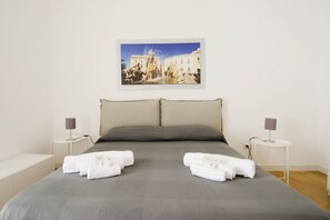 A neatly made bed with grey bedding and two pillows, a framed painting of a fountain above it, and two bedside tables with lamps. - La terrazza di Archimede Ortigia Holidays (Siracusa)