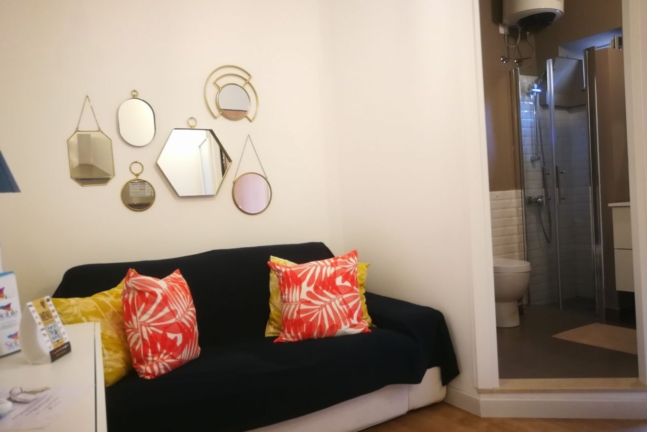 Ein Wohnzimmer mit einem dunkelgrauen Sofa, einem Couchtisch und einem an der Wand befestigten Spiegel. Durch eine Glastür ist eine Dusche zu sehen.