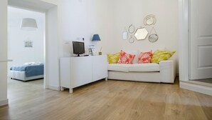 Ein modernes Wohnzimmer mit einem weißen Sofa, einem weißen Schrank und einem Fernseher. Dekorative Wandspiegel und eine Lampe ergänzen das Bild.
