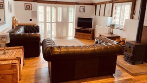 A cozy living room with a large leather sofa, a wooden coffee table, a television, and a wood-burning stove.
