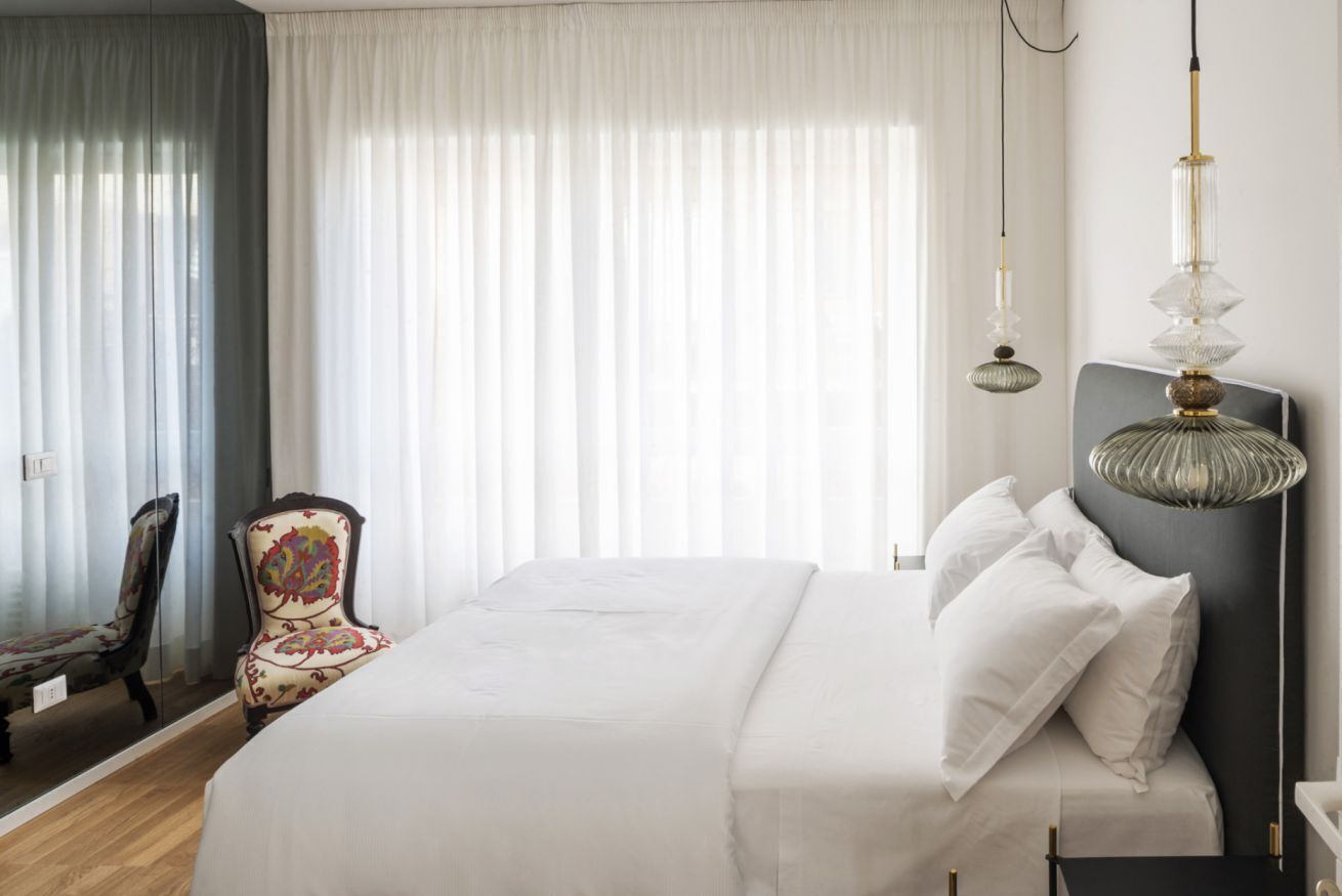 Ein ordentlich bezogenes Bett mit weißen Leinen, ein Sessel mit gemustertem Sitz und eine Pendelleuchte mit transparenter Kappe.