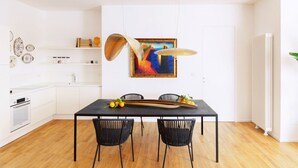 Eine moderne Küche mit weißen Schränken, einem schwarzen Esstisch und schwarzen Stühlen. An der Wand hängt ein farbenfrohes abstraktes Gemälde, und ein weißer Heizkörper steht auf dem Holzboden.