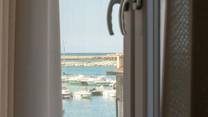 Blick durch ein Fenster auf einen Yachthafen mit vertäuten Booten, einem steinernen Wellenbrecher und einem wolkenlosen Himmel.