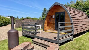 A wooden cabin with a curved roof, a small porch, and a grill area. - MegaPod 1 at Lee Wick Farm Glamping (Clacton on Sea)